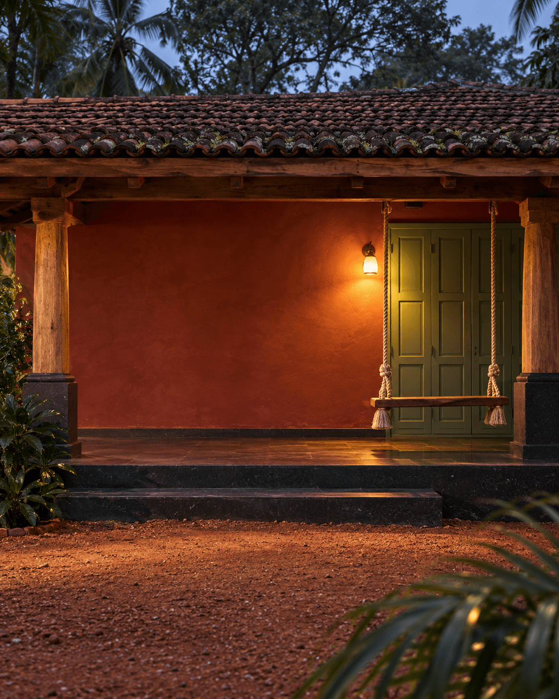 A red-oxide verandah at dusk with a rope swing, a lit lamp and green wooden doors.