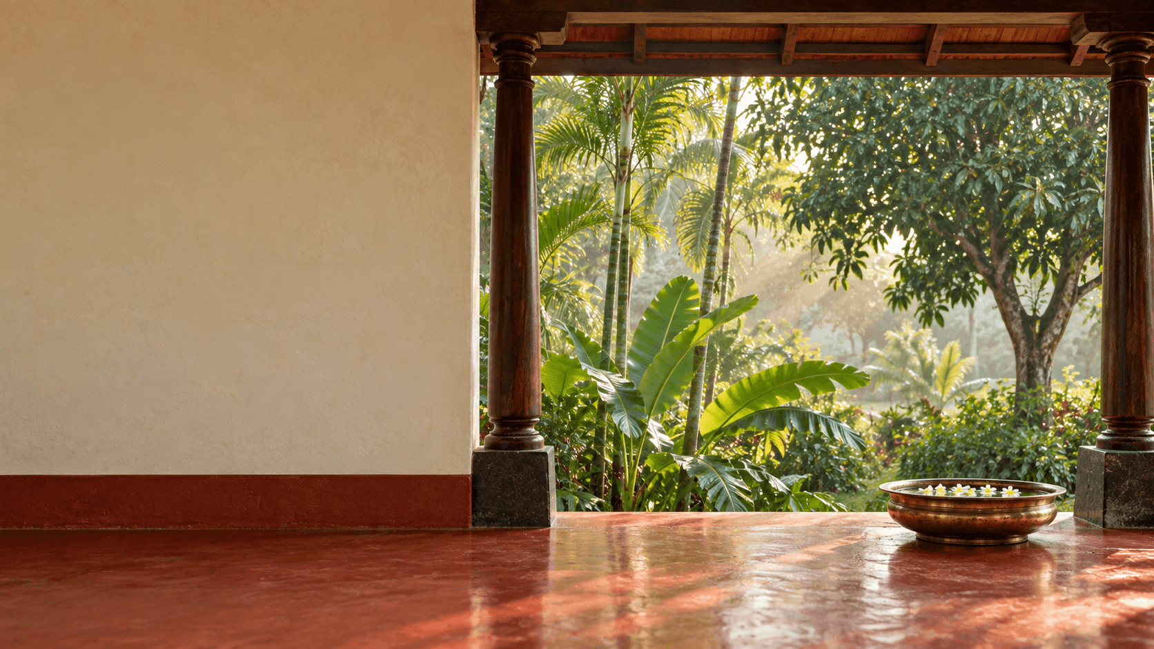 A cool morning verandah: an ivory wall, teak pillars, a brass uruli with floating flowers, and banana leaves and palms in the mist beyond.